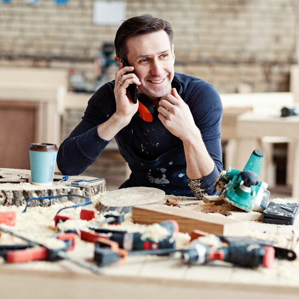 Portrait of young smiling carpenter talking on mobile phone at workbench surrounded by tools, wood slices and paper coffee cup